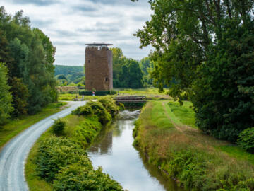 Een rustige natuurlandschap met een smalle waterloop, omringd door groen, met op de achtergrond een oude ronde bakstenen toren, de Maagdentoren.