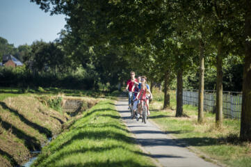 Drie fietsers achter elkaar fietsen op verharde weg langs beekje.