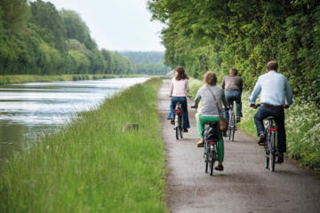 Vier mensen fietsen langs een pad, met veel natuur langs de kant, naast de Vaart.
