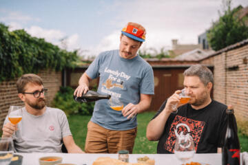 Drie mannen genieten in een tuin van een glas bier, terwijl één van hen inschenkt.