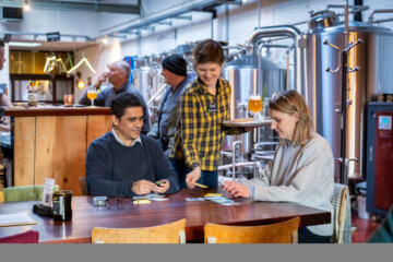 In een gezellige brouwerijomgeving zitten twee mensen aan een houten tafel een kaartspel te spelen terwijl een serveerster hen een kaart en een drankje aanreikt.