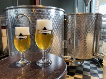 Twee volle glazen bier op en tafel met op de achtergrond twee brouwketels.